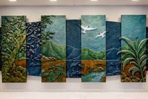 5’ x 10.5’ x 2.5” acrylic on hand carved Precision Board
In this Hawaiian mountain valley landscape, the scene is broken up into two levels symbolizing Day and Night, the Present and the Past. The panels of the day are vertical and geometric, lit by daylight, standing like skyscrapers in a modern city skyline. The panels of the night recede to the back, illuminated by the silver blue light of the full moon and shaped like an Ark carrying the present on its way to the future. Amidst the landscape are four native plants, Kalo, Ti, Kukui and Maile, that represent in ancient Hawaiian times essential qualities of Justice. There are also two animal spirits or aumakua, the Owl, always watchful, and the mated pair of Fairy Terns, sentinels of the City of Honolulu.
Created for the law offices of Goodsill, Anderson, Quinn & Stifel in downtown Honolulu, Hawai’i .
Completed February 2013