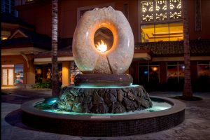 Aina … the sculpture hewn from granite the color of polished warm sand. It is a cycle of the elements of Earth, Air, Fire and Water, those very elements that continuously shape all that surrounds us. Inspired by the classic Hawaiian ring poi pounder, pohaku ku’i ai puka, this monumental ten ton arch floats on the surging waters that erupt around its three ton, black and red granite bowl or papa ku’iai and cascade down the lava rock he’eau that surrounds it. Cutting across the top of the ring is the carved profile of the islands surrounded by a moat of water that spills on either side down the roughly textured channels that flair to its base reminding us of Hawaii’s many stunning waterfalls. Within the center of the arch which defines the element of Air there is a central flowing well of Water that overflows on either side in textured flaring channels into the bowl. Emerging from the center of this well and framed by the arch of granite burns the element of Fire.
Commissioned by the Hilton Hawaiian Village in Waikiki, Hawai’i
Completed in December, 2008