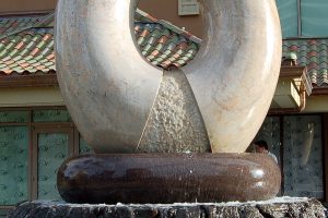Aina … the sculpture hewn from granite the color of polished warm sand. It is a cycle of the elements of Earth, Air, Fire and Water, those very elements that continuously shape all that surrounds us. Inspired by the classic Hawaiian ring poi pounder, pohaku ku’i ai puka, this monumental ten ton arch floats on the surging waters that erupt around its three ton, black and red granite bowl or papa ku’iai and cascade down the lava rock he’eau that surrounds it. Cutting across the top of the ring is the carved profile of the islands surrounded by a moat of water that spills on either side down the roughly textured channels that flair to its base reminding us of Hawaii’s many stunning waterfalls. Within the center of the arch which defines the element of Air there is a central flowing well of Water that overflows on either side in textured flaring channels into the bowl. Emerging from the center of this well and framed by the arch of granite burns the element of Fire.
Commissioned by the Hilton Hawaiian Village in Waikiki, Hawai’i
Completed in December, 2008