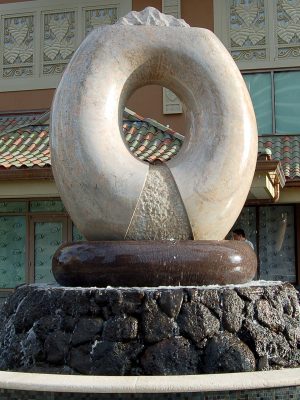 Aina … the sculpture hewn from granite the color of polished warm sand. It is a cycle of the elements of Earth, Air, Fire and Water, those very elements that continuously shape all that surrounds us. Inspired by the classic Hawaiian ring poi pounder, pohaku ku’i ai puka, this monumental ten ton arch floats on the surging waters that erupt around its three ton, black and red granite bowl or papa ku’iai and cascade down the lava rock he’eau that surrounds it. Cutting across the top of the ring is the carved profile of the islands surrounded by a moat of water that spills on either side down the roughly textured channels that flair to its base reminding us of Hawaii’s many stunning waterfalls. Within the center of the arch which defines the element of Air there is a central flowing well of Water that overflows on either side in textured flaring channels into the bowl. Emerging from the center of this well and framed by the arch of granite burns the element of Fire.
Commissioned by the Hilton Hawaiian Village in Waikiki, Hawai’i
Completed in December, 2008