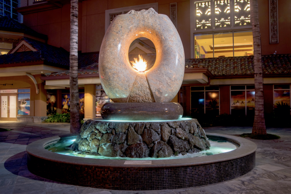 Hilton Grand Vacation Waikiki
Aina … the sculpture hewn from granite the color of polished warm sand. It is a cycle of the elements of Earth, Air, Fire and Water, those very elements that continuously shape all that surrounds us. Inspired by the classic Hawaiian ring poi pounder, pohaku ku’i ai puka, this monumental ten ton arch floats on the surging waters that erupt around its three ton, black and red granite bowl or papa ku’iai and cascade down the lava rock he’eau that surrounds it. Cutting across the top of the ring is the carved profile of the islands surrounded by a moat of water that spills on either side down the roughly textured channels that flair to its base reminding us of Hawaii’s many stunning waterfalls. Within the center of the arch which defines the element of Air there is a central flowing well of Water that overflows on either side in textured flaring channels into the bowl. Emerging from the center of this well and framed by the arch of granite burns the element of Fire.
Commissioned by the Hilton Hawaiian Village in Waikiki, Hawai’i
Completed in December, 2008